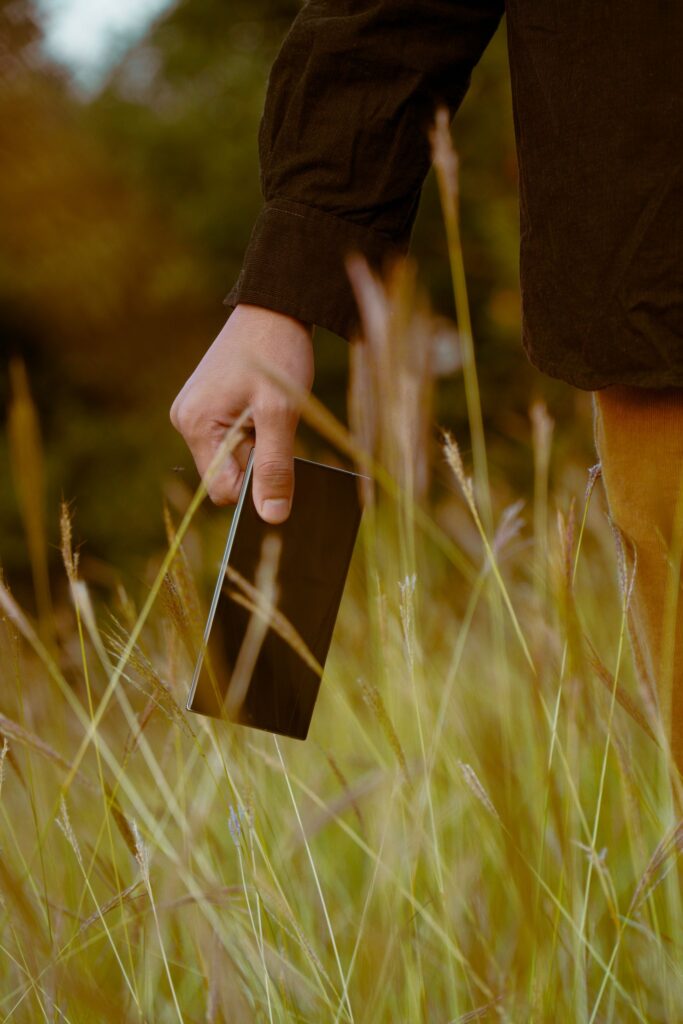 現在の画像: A close-up shot of a person holding a mobile phone in a field of tall grass.