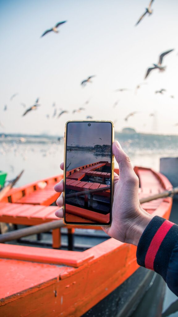 現在の画像: A hand holding a smartphone capturing birds flying over a lake with boats in the background.