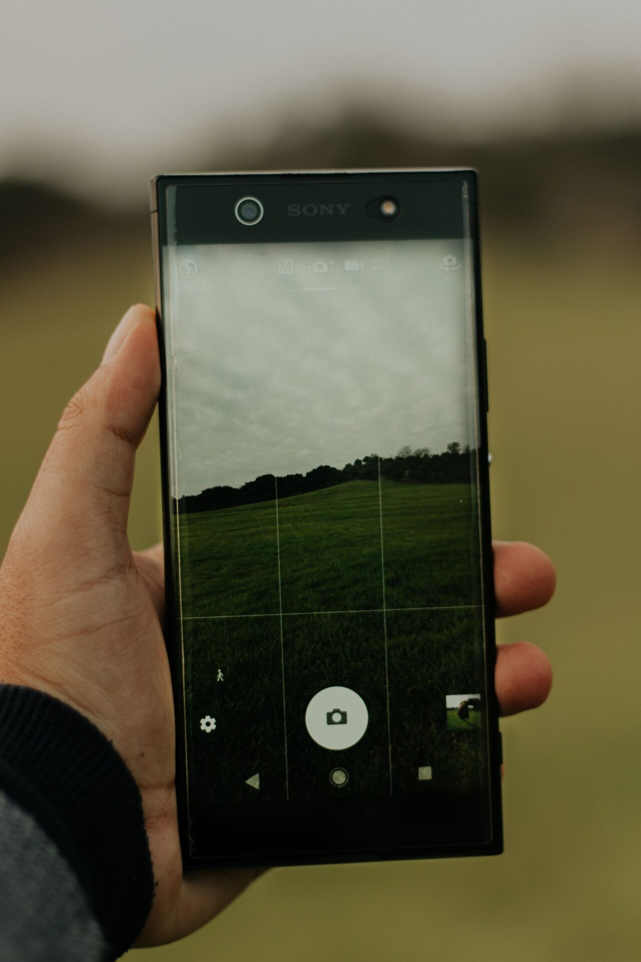 A hand holds a smartphone showing a camera view of a grassy field.