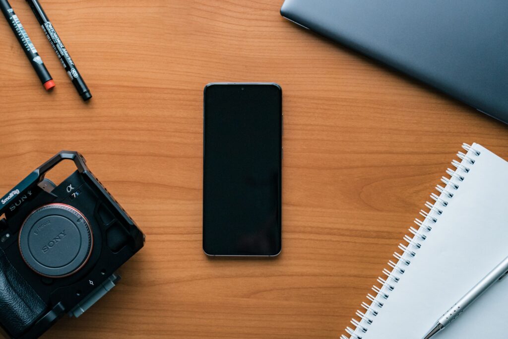 現在の画像: A neat flat lay of a modern workspace featuring a smartphone, camera, pens, and notebook.
