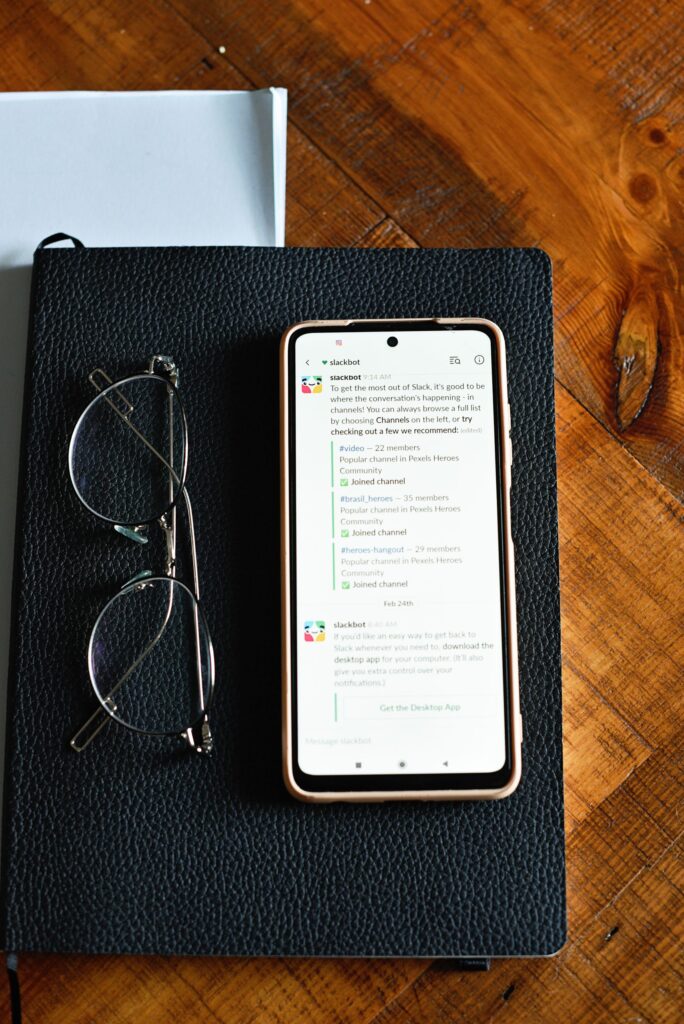 現在の画像: A smartphone displaying text is placed on a notepad alongside eyeglasses, on a wooden table.