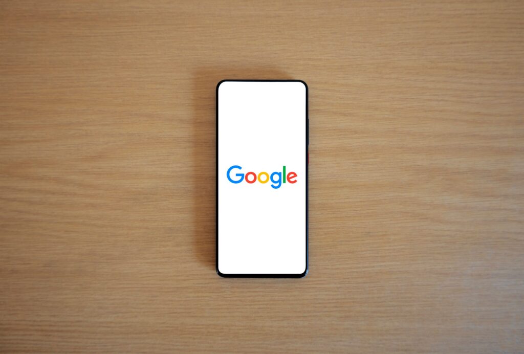 現在の画像: A smartphone displaying the Google homepage on a wooden surface, viewed from above.