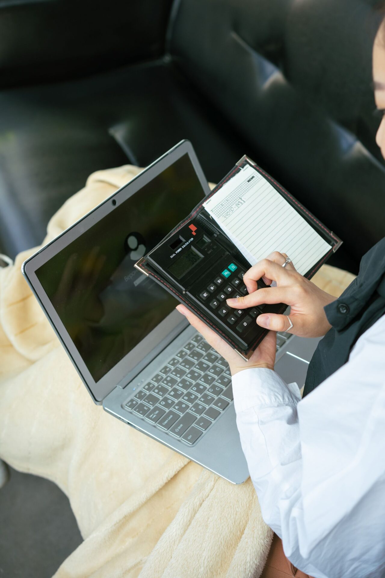 A woman using a calculator while working from a laptop on a couch, showcasing multitasking.