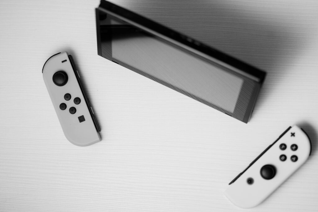 現在の画像: Black and white minimalist flat lay of Nintendo Switch console with game controllers on a white surface.