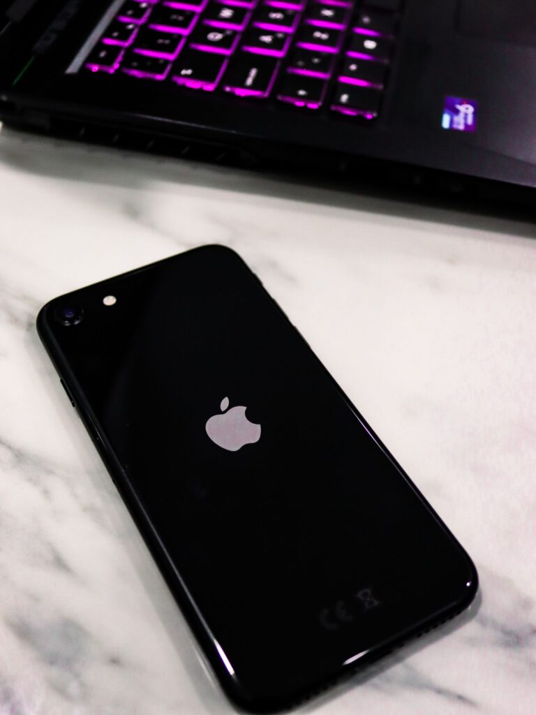 Close-up of a black smartphone with an apple logo on a marble surface next to a laptop with illuminated keys.