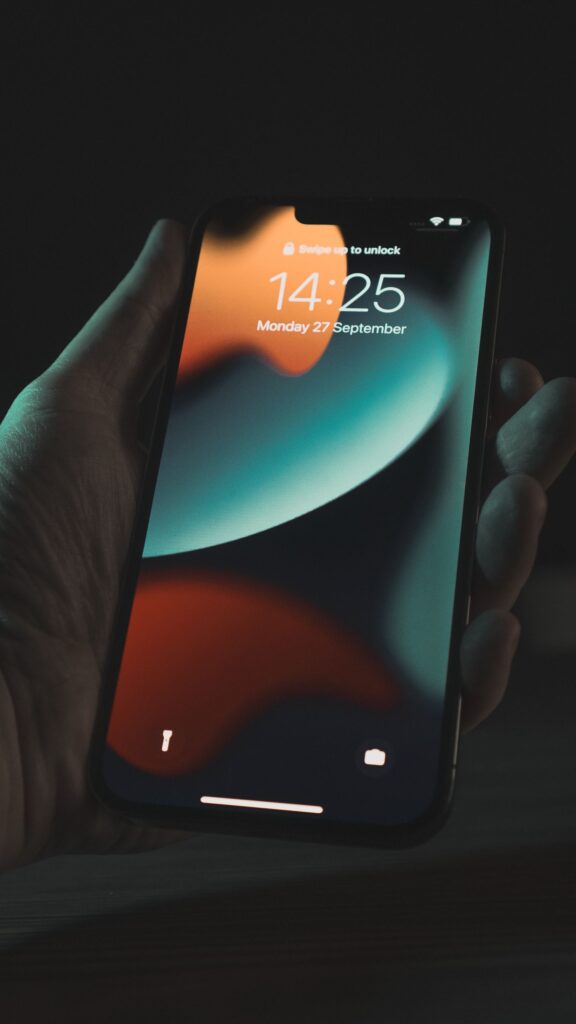 現在の画像: Close-up of a hand holding a smartphone displaying the lockscreen and time in a dimly lit setting.