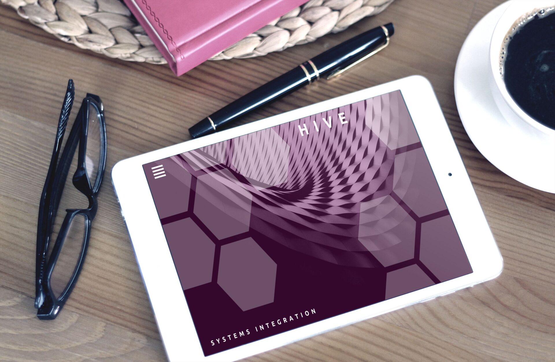 Flat lay of a modern workspace featuring a tablet displaying a screen, glasses, coffee, and a pen on a wooden desk.