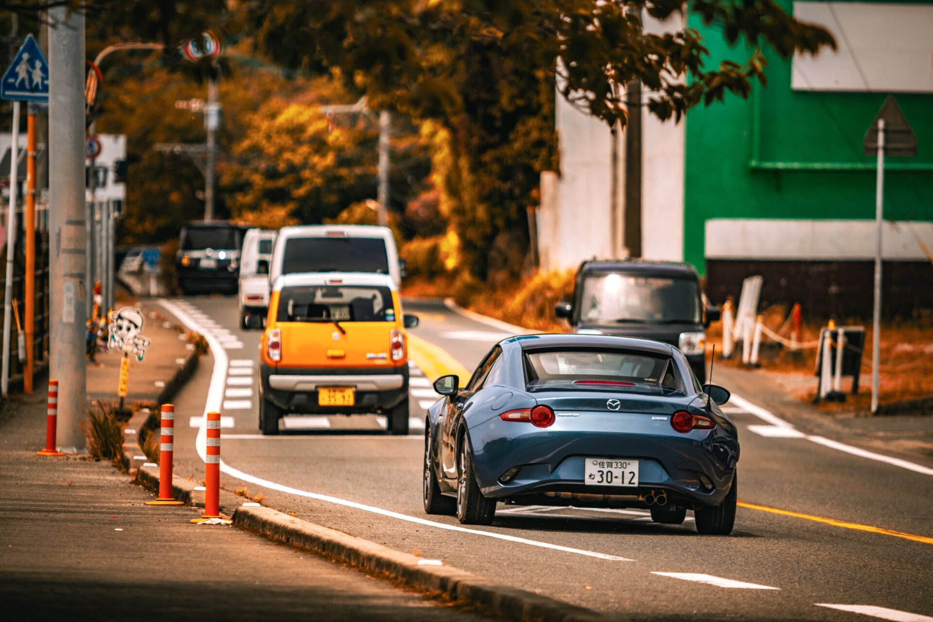 Mazda MX-5 car driving on an urban street surrounded by autumn foliage and city elements.