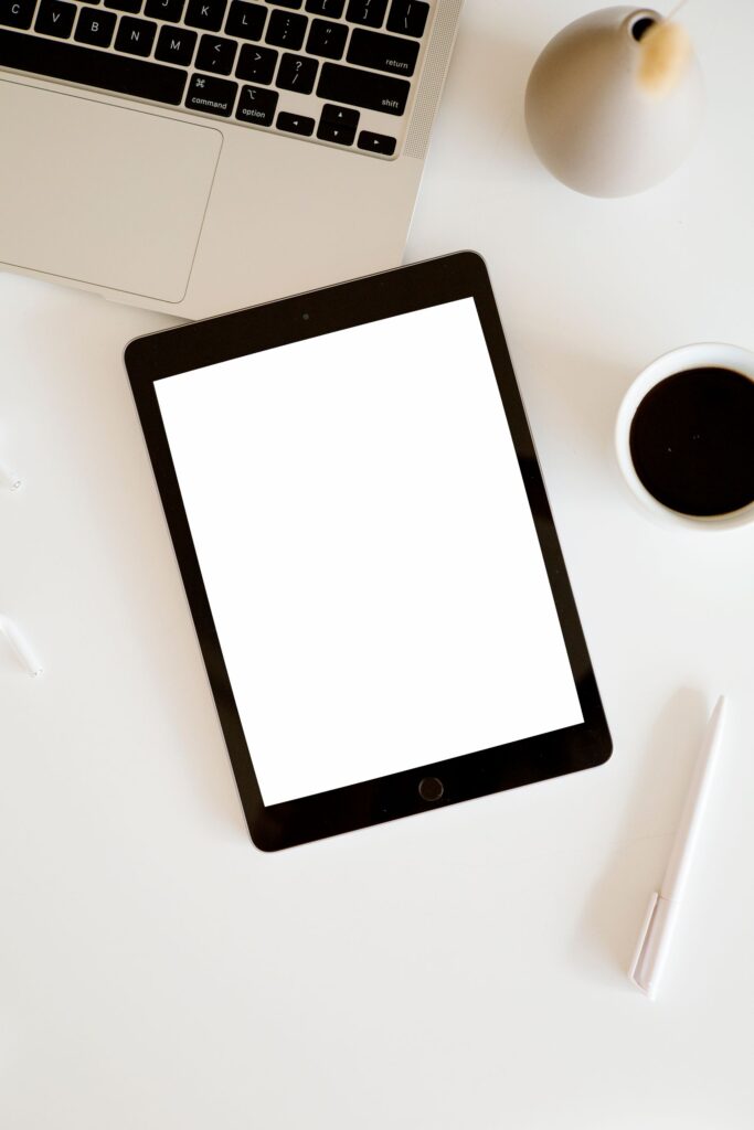 現在の画像: Minimalist flat lay with a tablet, black coffee, laptop, and pen on a white surface.