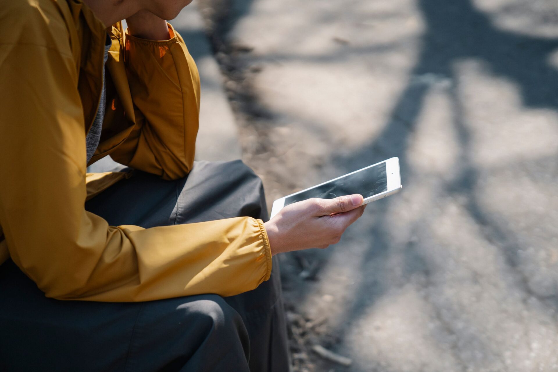 Side view of crop anonymous person in yellow jacket sitting on pavement and holding tablet