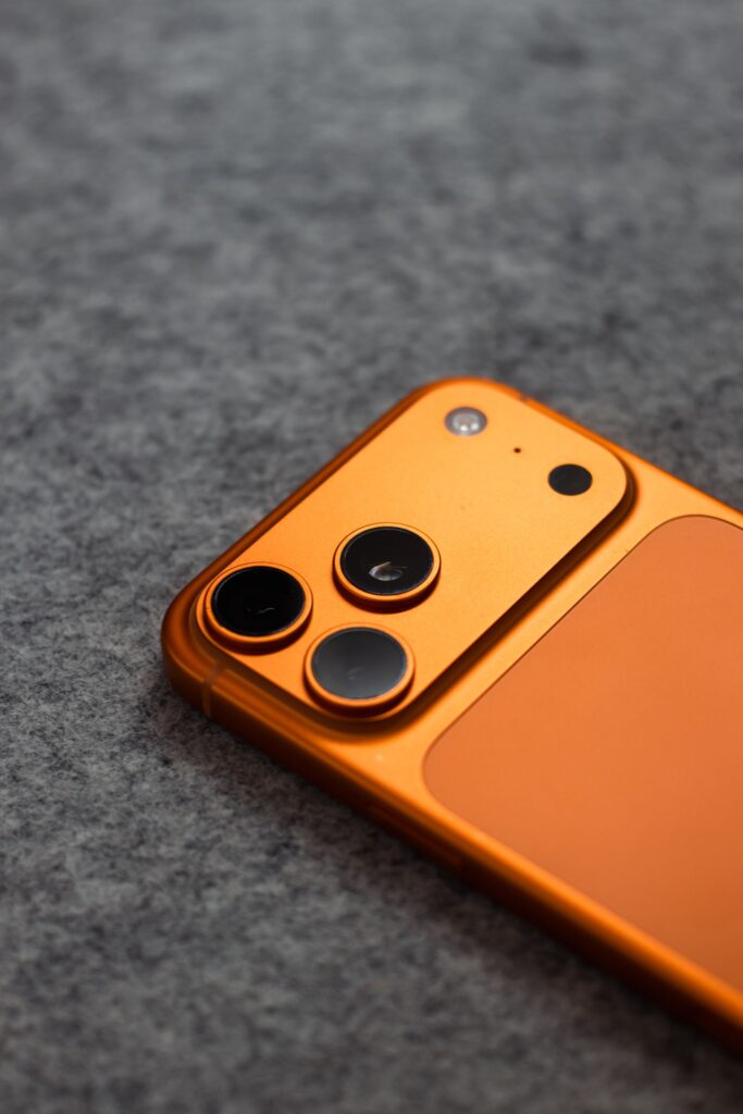 現在の画像: Stylish orange smartphone with triple camera lens on a gray fabric background, perfect for tech themes.